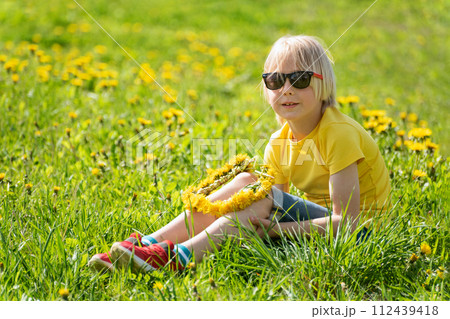 Fair-haired little child boy in yellow t-shirt sits on green grass lawn. Kid weaves wreath of dandelions sitting in field. 112439418