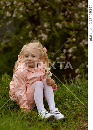 Fair-haired girl in pink dress sitting near blooming tree and looking in the camera. Cute child girl in the flowering garden. Vertical frame. 112439544