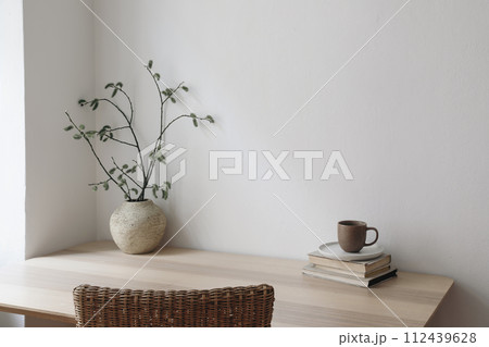 Scandinavian living room interior. Cup of coffee, books on wooden table. Willow tree branches, catkins in vase. Rattan chair. Elegant Scandi working space, home office. Blank beige wall background 112439628