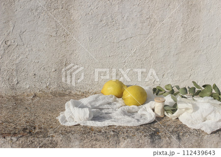 Wedding still life. Fresh lemons fruit, eucalyptus tree branches, ribbon and muslin textile in sunlight. White shabby old textured wall background. Vacation concept. Mediterranean summer banner. 112439643