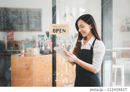 Beautiful young barista woman in apron holding order paper and standing in front of the door of cafe with open sign board. Business owner startup concept. Beautiful young barista woman in apron holding order paper and standing in front of the door of cafe with open sign board. Business owner startup concept. 112439827