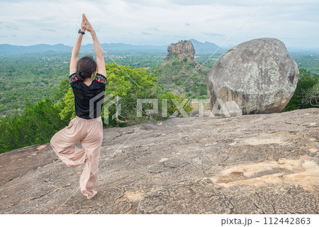 Back view of young woman doing Yoga on top of Pidurangala rock and looking to Sigiriya rock an iconic tourist destination and one of UNESCO world heritage site in Sri Lanka. Back view of young woman doing Yoga on top of Pidurangala rock and looking to Sigiriya rock an iconic tourist destination and one of UNESCO world heritage site in Sri Lanka. 112442863