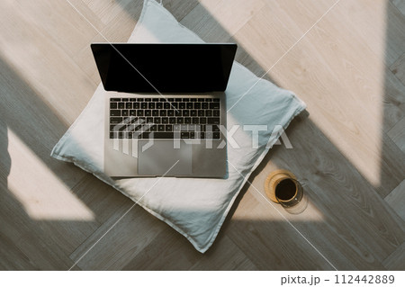 Photo of laptop showing black empty screen putting on white pillow, on wooden floor in cozy room with black coffee cup. 112442889