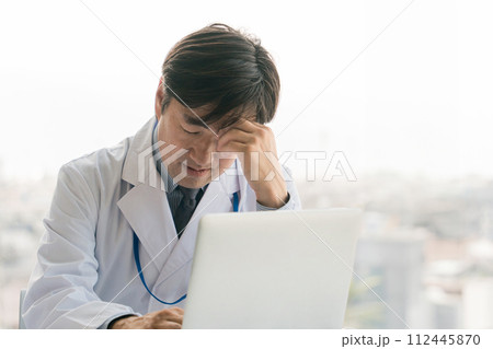 パソコンの前で悩む・考える・困る男性医師（眼精疲労・過労・疲労・多忙） 112445870