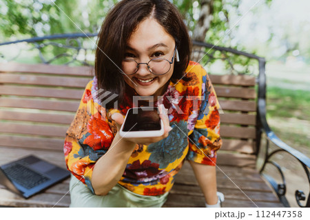 Happy woman sitting on a bench in a green park, emotionally talking on her phone. Smiling freelancer wearing a colorful blouse, animatedly chatting on her smartphone outdoor. Happy woman sitting on a bench in a green park, emotionally talking on her phone. Smiling freelancer wearing a colorful blouse, animatedly chatting on her smartphone outdoor. 112447358