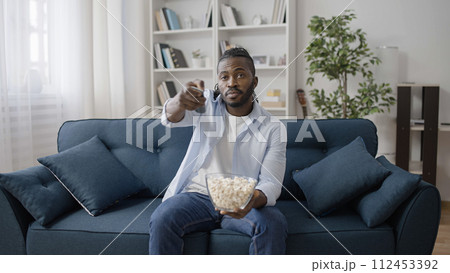 African American man sitting on sofa, watching movie on TV and eating popcorn 112453392