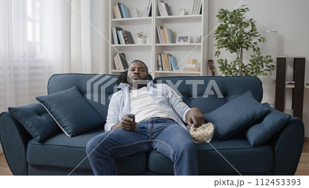 Carefree African American man eating popcorn, lying on sofa and watching TV Carefree African American man eating popcorn, lying on sofa and watching TV 112453393