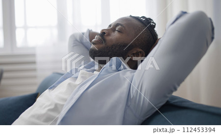 Tired African American man sitting back and relaxing on sofa at home after work day 112453394