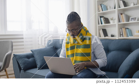 African American wrapped in scarf typing on laptop at home, working while ill 112453399