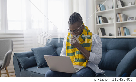 African American IT specialist in scarf coughing, working on laptop, having flu African American IT specialist in scarf coughing, working on laptop, having flu 112453400
