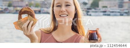 BANNER, LONG FORMAT Woman in Istanbul having breakfast with Simit and a glass of Turkish tea. Glass of Turkish tea and bagel Simit against golden horn bay in Istanbul, Turkey. Turkiye 112454644