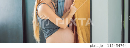 BANNER, LONG FORMAT A pregnant woman has nothing to wear. A pregnant woman stands in front of a closet with clothes and does not know what to wear because the clothes do not fit on her 112454764