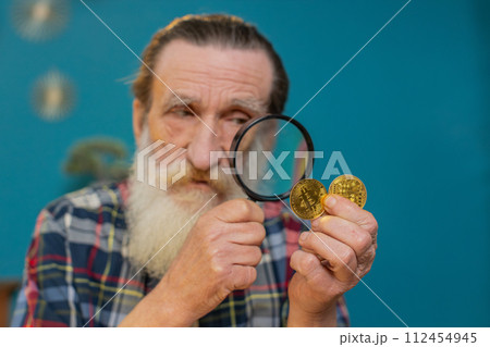 Surprise senior mature man examining golden bitcoins BTC with magnifying glass sitting at table Surprise senior mature man examining golden bitcoins BTC with magnifying glass sitting at table 112454945