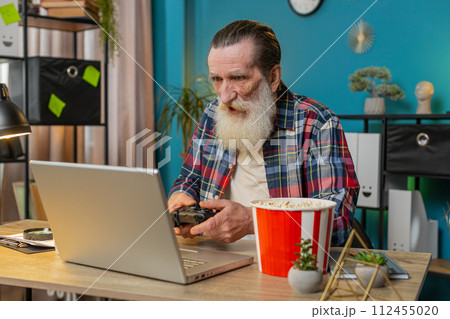 Excited happy senior man celebrating victory in play video game on laptop using controller joystick 112455020