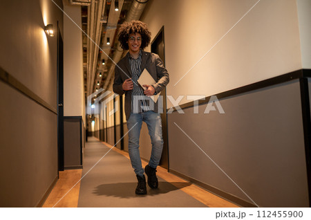 Curly-haired thin man with laptop walking in a hotel corridor 112455900