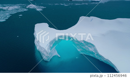 Giant Antarctica glacier arch aerial panorama. Snow covered iceberg with melted hole inside floating blue ocean water. Polar climate change. Arctic winter landscape, global warming problem. Drone Giant Antarctica glacier arch aerial panorama. Snow covered iceberg with melted hole inside floating blue ocean water. Polar climate change. Arctic winter landscape, global warming problem. Drone 112456946