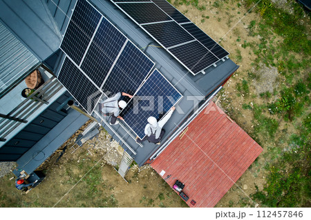 Installers building photovoltaic solar module station on roof of house. Men electricians in helmets installing solar panel system outdoors. Concept of alternative and renewable energy. Aerial view. Installers building photovoltaic solar module station on roof of house. Men electricians in helmets installing solar panel system outdoors. Concept of alternative and renewable energy. Aerial view. 112457846