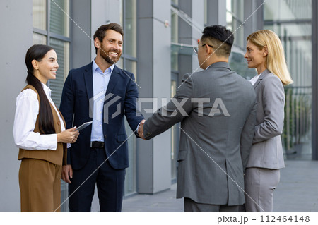 Business meeting of interracial businessmen. Men and women stand outside an office center and shake hands as a sign of getting to know each other and making a deal. Business meeting of interracial businessmen. Men and women stand outside an office center and shake hands as a sign of getting to know each other and making a deal. 112464148