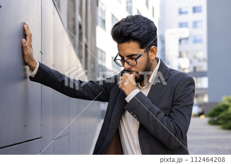 A young Indian man in a business suit is standing near the office, leaning his hand on the wall and suffering from a strong cough, covering his mouth with his hand. 112464208