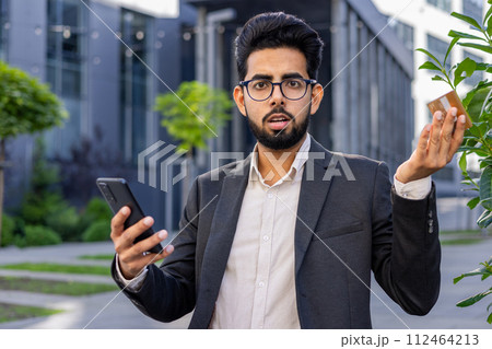 Portrait of a worried young Indian male businessman in a business suit sitting on a bench near the office, holding a credit card and a phone, looking disappointedly at the camera and spreading hands. 112464213