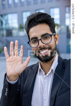 Vertical close-up photo of a young smiling Indian man in a suit and glasses talking to the camera on a video call, waving and greeting. 112464247