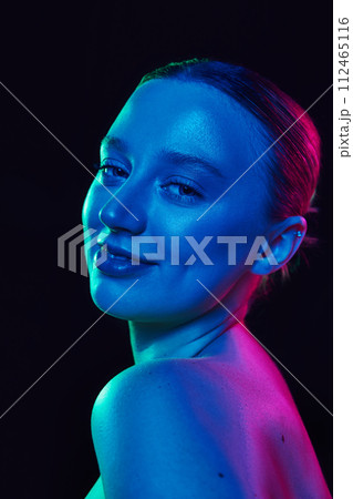Close of young attractive woman looking at camera with smile in neon light against black studio background. Model with well-kept glowing skin. 112465116