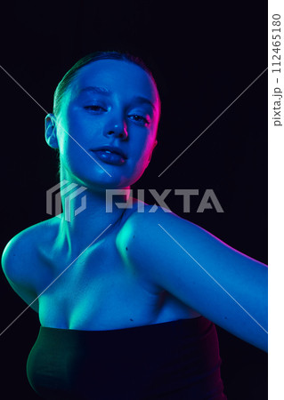 Portrait of young attractive woman in top looking at camera confidently in neon light against black studio background. Portrait of young attractive woman in top looking at camera confidently in neon light against black studio background. 112465180