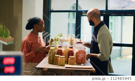 Owner recommends bulk products for woman, presenting variety of chemicals free grains and cooking ingredients stored in reusable jars. Storekeeper explains ecological production process to client. 112465945