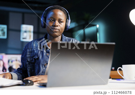 Focused black woman wearing wireless headphones using laptop and writing notes. African american female student with headset watching online webinar, listening to audio course on personal computer. 112466078