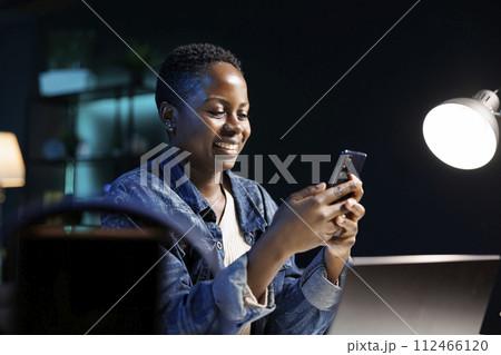Cheerful black woman utilizing modern technology, taking break from laptop and looking at social media on mobile device. African american lady surfing the net on cellphone, online communication. 112466120
