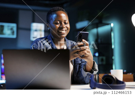Cheerful female college student using mobile device to engage with friends on social media platforms. African american lady using cell phone to surf the web while sitting at desk in her home office. Cheerful female college student using mobile device to engage with friends on social media platforms. African american lady using cell phone to surf the web while sitting at desk in her home office. 112466153