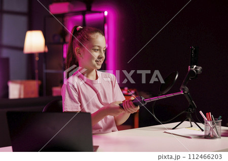 Cute girl filming room tour inside home studio using smartphone on selfie stick. Little kid doing video presenting her neon lit apartment to interested children audiences Cute girl filming room tour inside home studio using smartphone on selfie stick. Little kid doing video presenting her neon lit apartment to interested children audiences 112466203