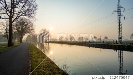 Misty Canal Pathway at Sunrise with Reflective Water and Power Lines Misty Canal Pathway at Sunrise with Reflective Water and Power Lines 112470047