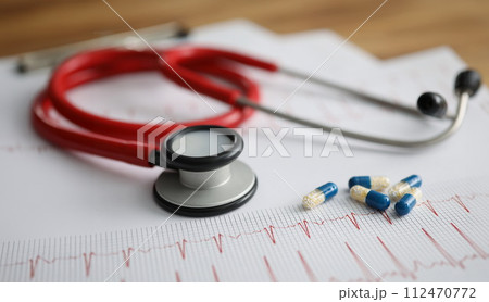 Medical capsules and red stethoscope lying on electrocardiogram closeup Medical capsules and red stethoscope lying on electrocardiogram closeup 112470772