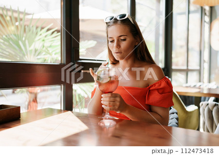 woman in a cafe seated, holding beverage. Dressed in an orange dress with her hair down. Bright interior, large windows allow natural light. Beverage consumption for refreshment. woman in a cafe seated, holding beverage. Dressed in an orange dress with her hair down. Bright interior, large windows allow natural light. Beverage consumption for refreshment. 112473418