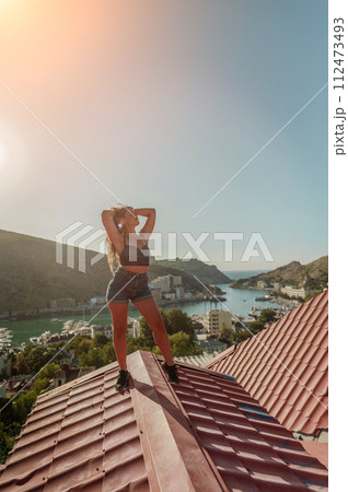 Woman standing on rooftop, enjoys town view and sea mountains. Peaceful rooftop relaxation. Below her, there is a town with several boats visible in the water. Rooftop vantage point. 112473493