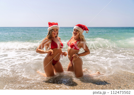 Women Santa hats ocean play. Seaside, beach daytime, enjoying beach fun. Two women in red swimsuits and Santa hats are enjoying themselves in the ocean waves. 112473502