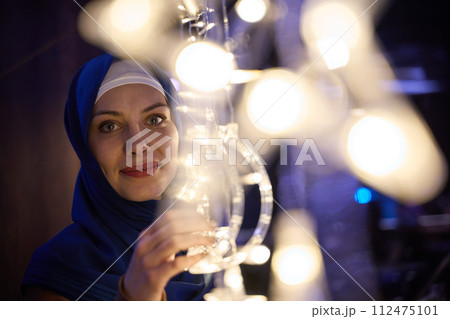 In a modern restaurant ambiance, a woman in a hijab captures a selfie beside glowing lights, showcasing contemporary style and cultural diversity in a trendy urban setting. 112475101