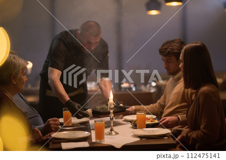 In a restaurant setting, a professional chef presents a sizzling steak cooked over an open flame, while an European Muslim family eagerly awaits their iftar meal during the holy month of Ramadan 112475141
