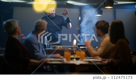 In a restaurant setting, a professional chef presents a sizzling steak cooked over an open flame, while an European Muslim family eagerly awaits their iftar meal during the holy month of Ramadan 112475173