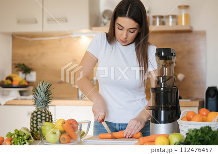 Attractive young woman cutting carrot in kitchen. Female preparing vegetables and fruits for juice. Home made fresh. Juicer Attractive young woman cutting carrot in kitchen. Female preparing vegetables and fruits for juice. Home made fresh. Juicer 112476457