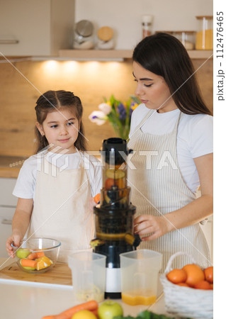 Adorable five year old girl putting with her mom fruits into juicer. Two females into aprons. 112476465