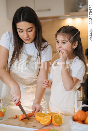 Attractive young mom slice orange with her adorable five year old daughter. Family preparing vegetables and fruits for Juicer. Fresh homemade juice 112476474