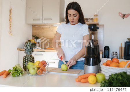 Attractive young woman cutting apple in kitchen. Female preparing vegetables and fruits for juice. Home made fresh. Juicer Attractive young woman cutting apple in kitchen. Female preparing vegetables and fruits for juice. Home made fresh. Juicer 112476475