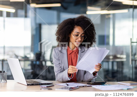 A focused professional woman holds and reviews paperwork at her organized office workstation, displaying productivity and concentration. 112476899