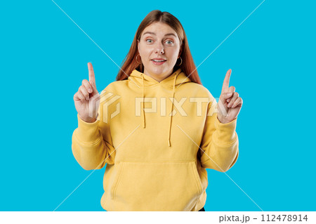 Cheerful happy female model pointing fingers up to shop promotion banner, smiling camera, studio portrait. 30s Woman pointing fingers at copy space isolated over blue background, surprised face. 112478914
