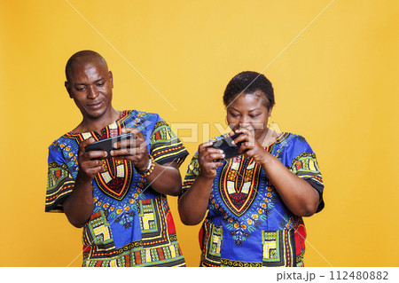 Cheerful african american wife and husband pair enjoying mobile game competition. Smiling couple playing entertaining game on smartphones, sharing fun leisure activity together 112480882