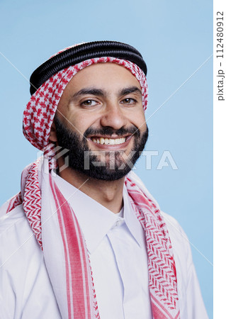 Smiling arab man wearing traditional headscarf looking at camera with cheerful face expression. Joyful muslim person in ghutra headdress posing for studio portrait on blue background 112480912