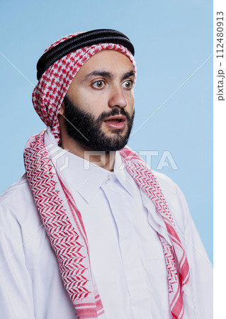 Shocked muslim man dressed in traditional headscarf looking away with wide eyes and open mouth. Person wearing arab ghutra and thobe clothes while posing with surprised face expression 112480913