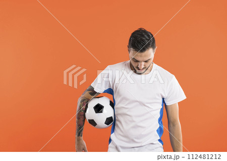 Soccer player looking down and holding a ball with one arm isolated on orange background 112481212